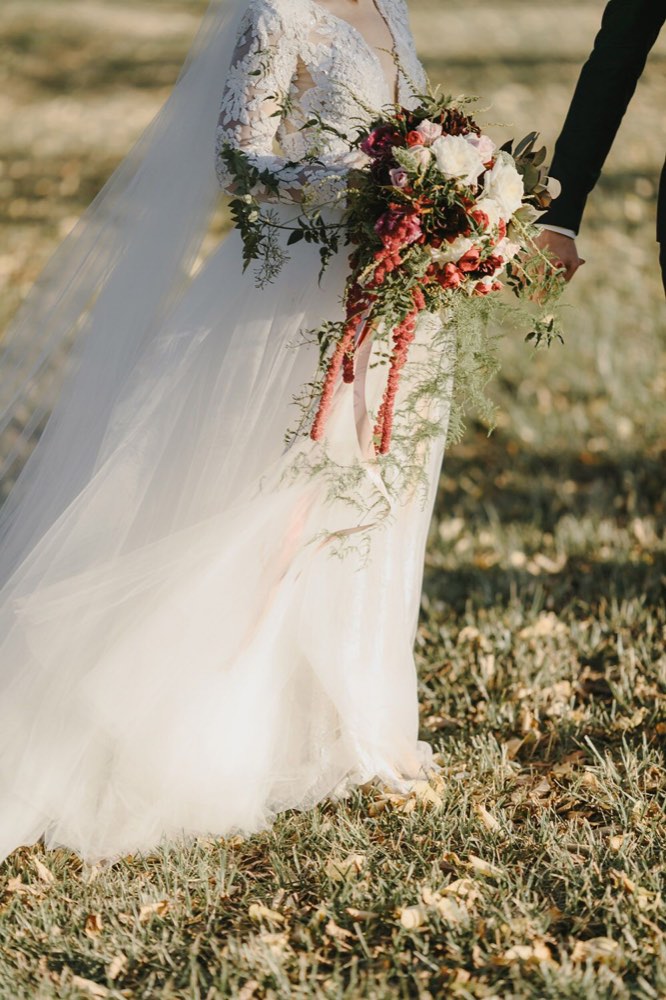 Trailing bridal bouquet with deep red blooms and ribbon, held beside a lace gown