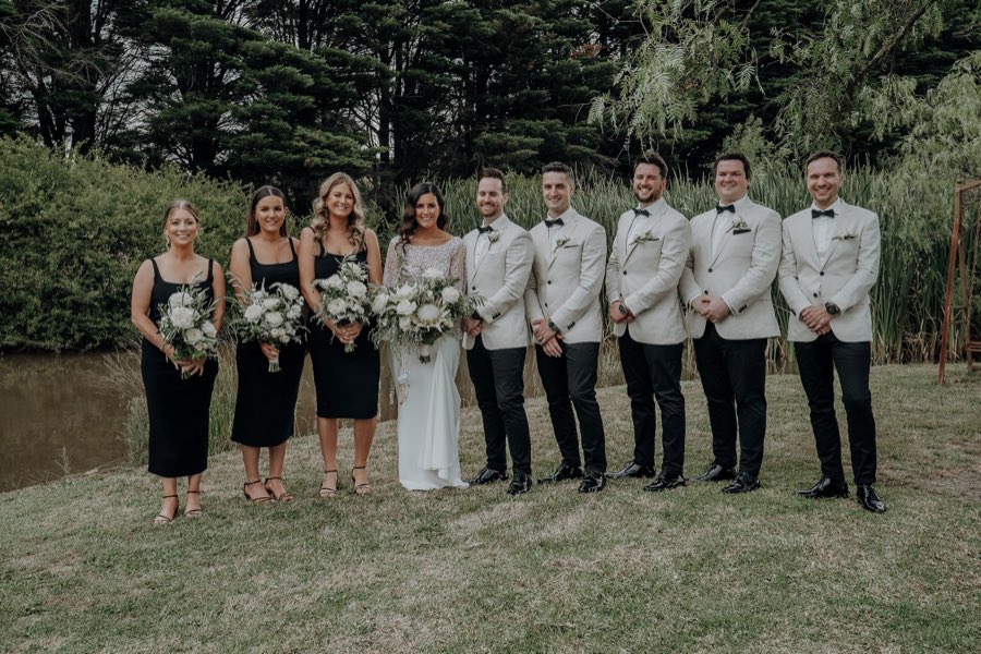 Black tie bridal party on a lawn beside water, holding white and green bouquets